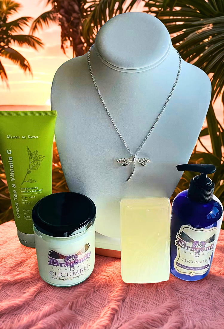 A curated wellness gift set arranged on a pink textured blanket in front of a silver dragonfly necklace. The set includes Green Tea & Vitamin C face wash , a candle, soap and lotion.