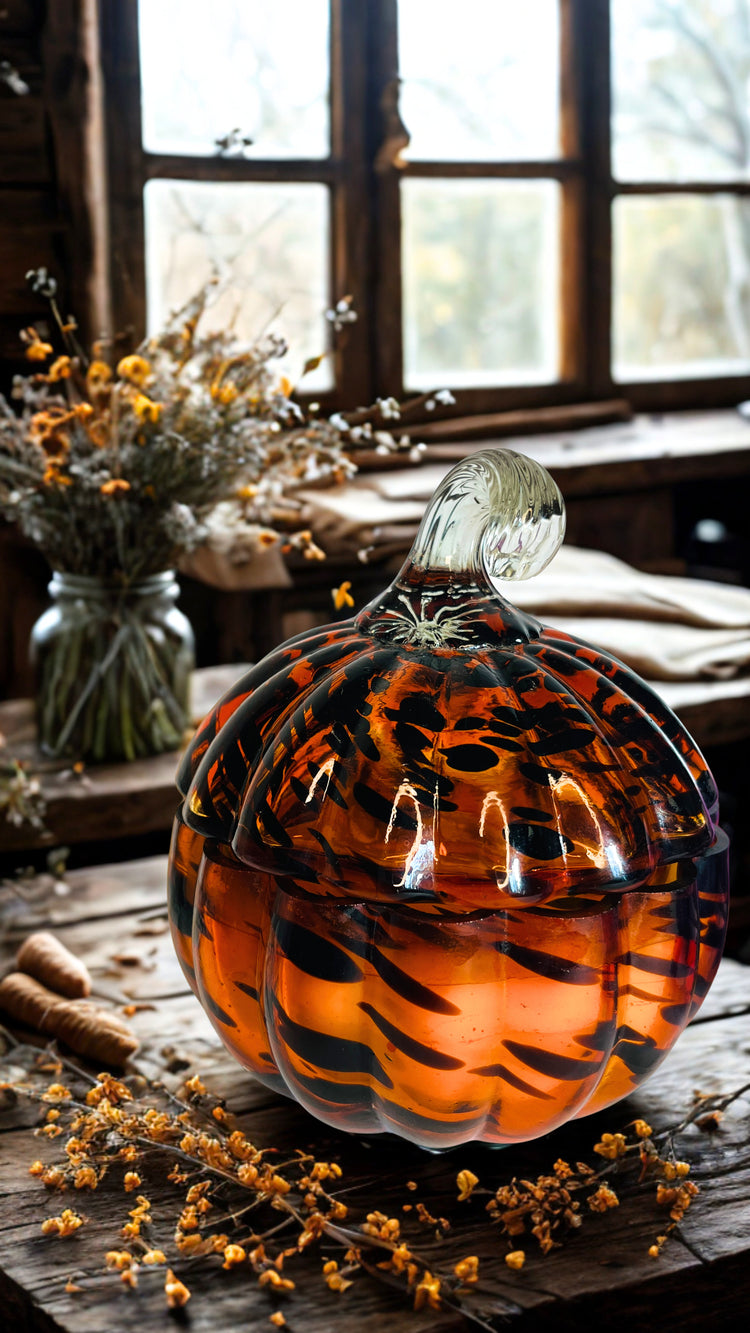 A decorative glass pumpkin candle with a ribbed pumpkin shape, filled with amber-orange accents with dark tiger-stripe patterns, set on a rustic wooden table with dried flowers and a window in the background.