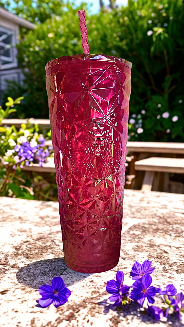A red Starbucks tumbler with a diamond-cut pattern and a pink striped straw, set outdoors on a stone surface with purple flowers and green foliage in the background.