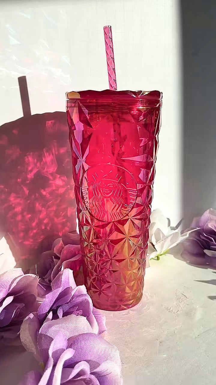 A textured, faceted pink Starbucks tumbler with a matching pink striped straw, set against a white background with decorative purple flowers and a soft shadow.