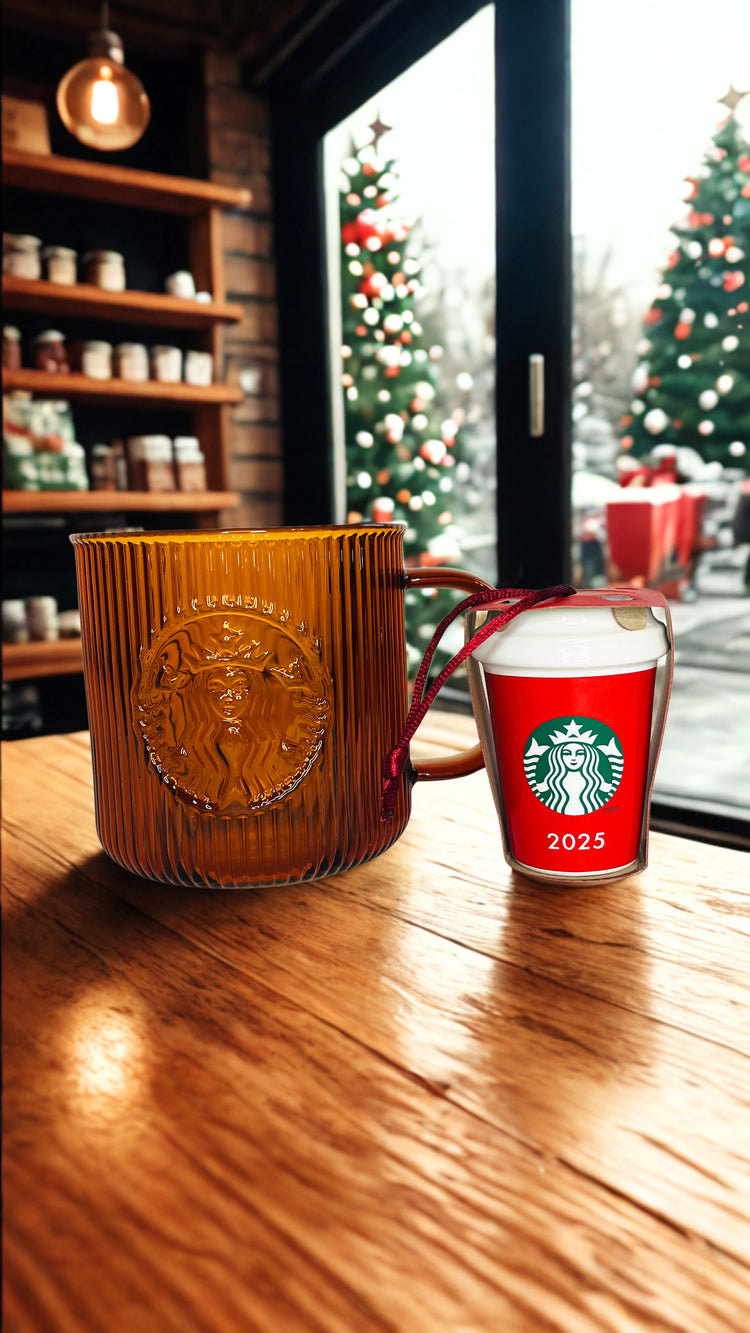 Starbucks mug and red ornament with 2025 design on a wooden table, Christmas trees visible through window.