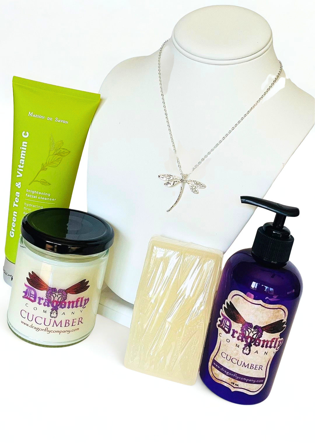 A product bundle featuring a silver dragonfly necklace, a green tube of brightening facial cleanser, a jar of Dragonfly Cucumber candle, a wrapped bar of soap, and a purple pump bottle of Dragonfly  Cucumber lotion all on a white background.
