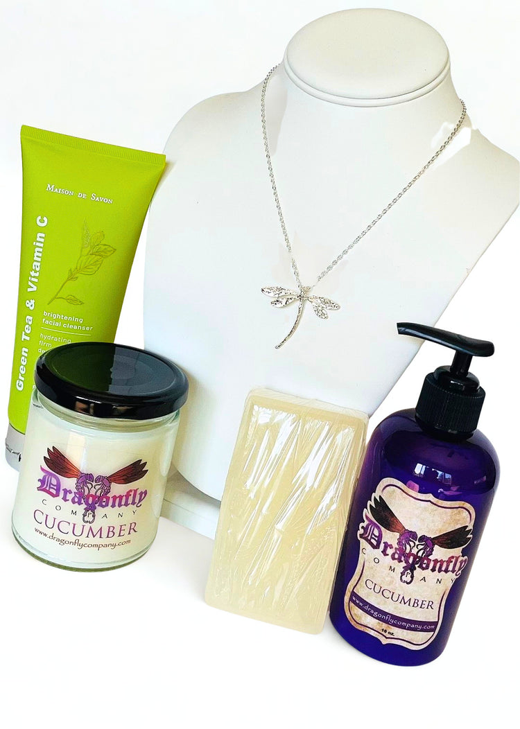 A product bundle featuring a silver dragonfly necklace, a green tube of brightening facial cleanser, a jar of Dragonfly Cucumber candle, a wrapped bar of soap, and a purple pump bottle of Dragonfly  Cucumber lotion all on a white background.