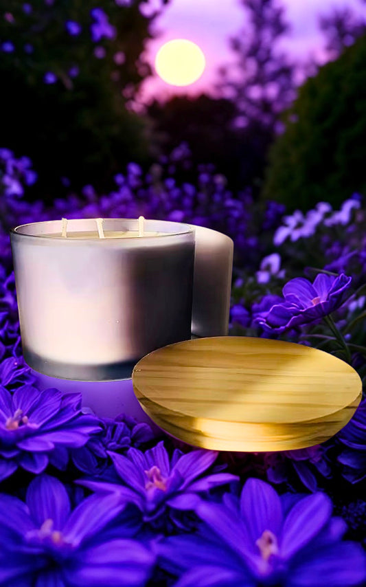 A lit  candle in a clear glass jar sits on a bed of purple flowers, with a round wooden lid placed beside it. The background is a soft-focus outdoor garden scene at dusk with purple foliage and a glowing sun.