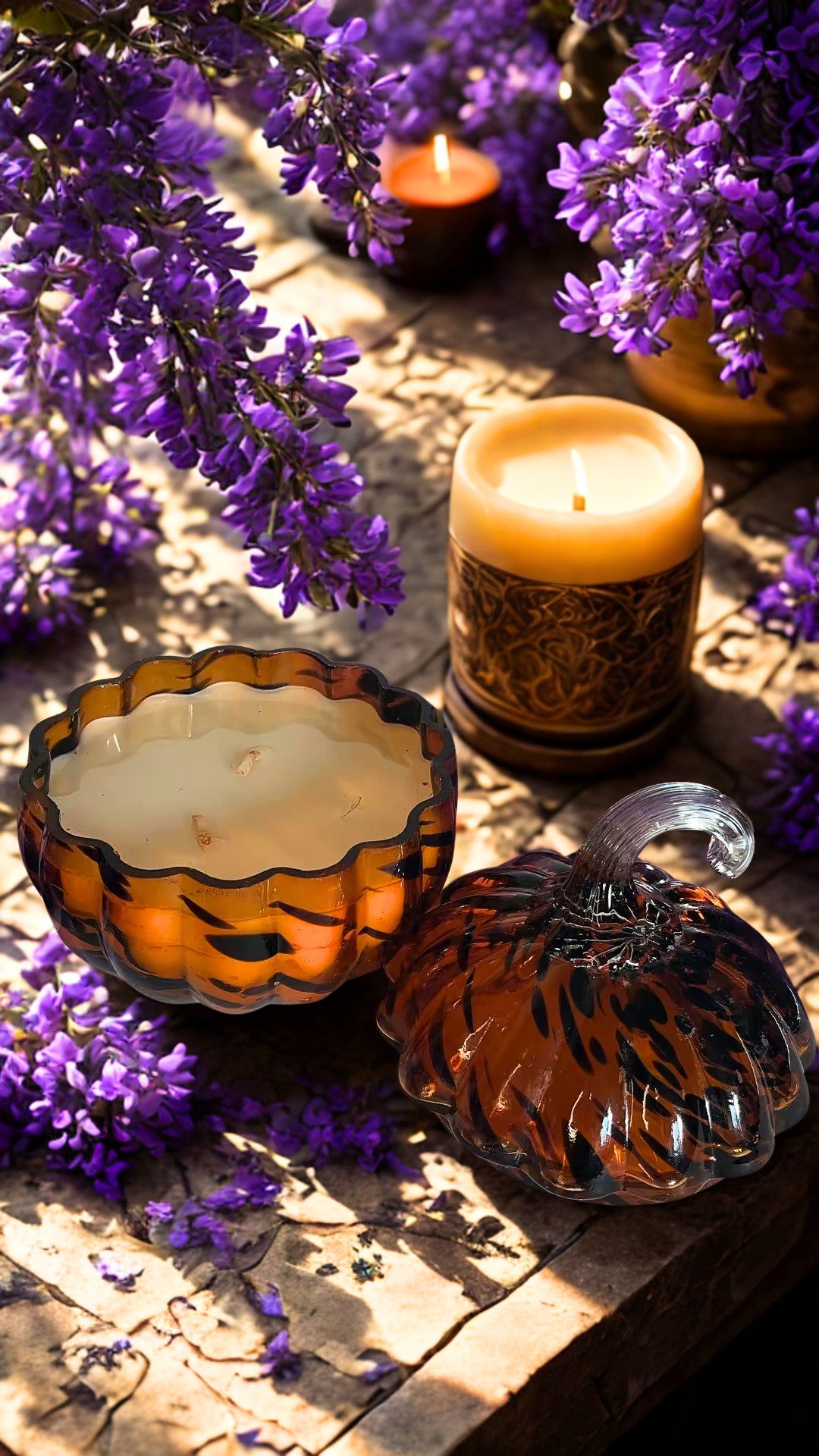 Decorative candles and glass containers arranged on a rustic wooden surface with scattered purple flowers. and a clear glass pumpkin-shaped container candle with an amber swirl pattern.