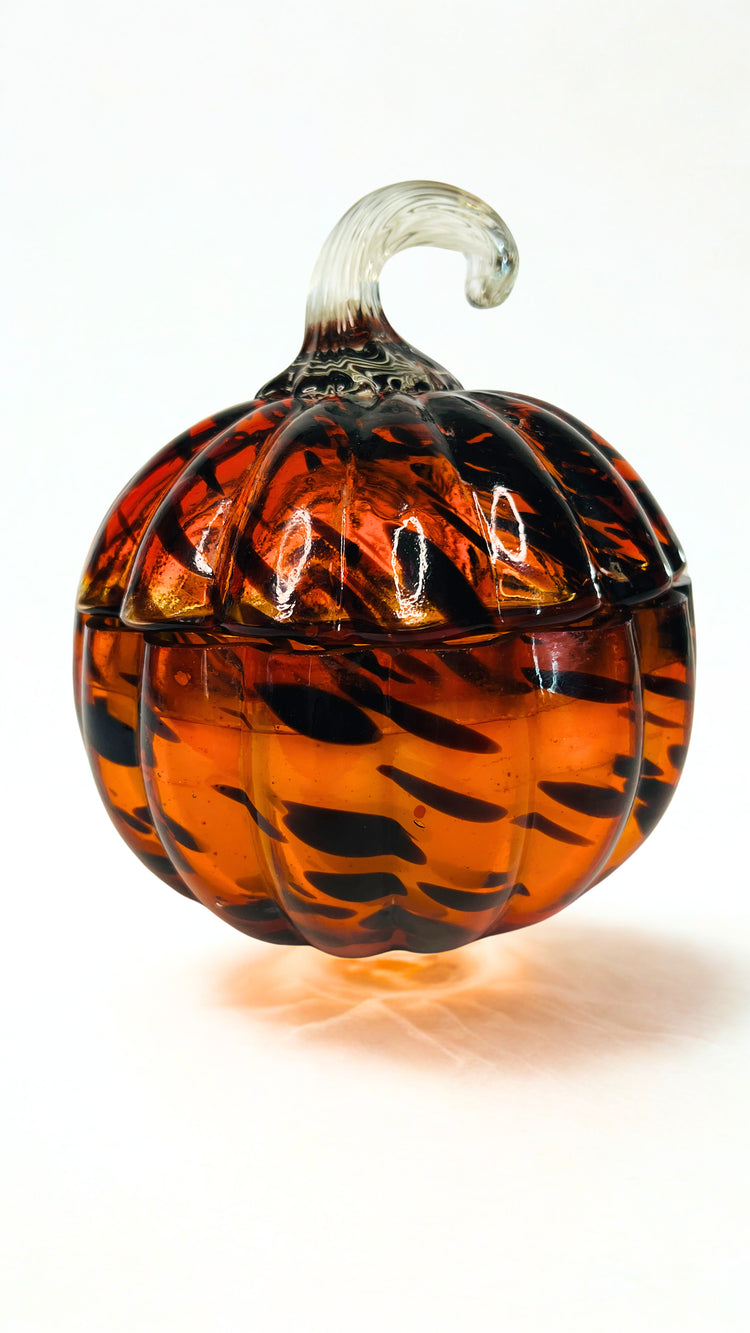 A decorative glass pumpkin container with a lid, featuring a swirled amber orange and black pattern, a clear glass stem, and a small clear glass base, set against a plain white background.