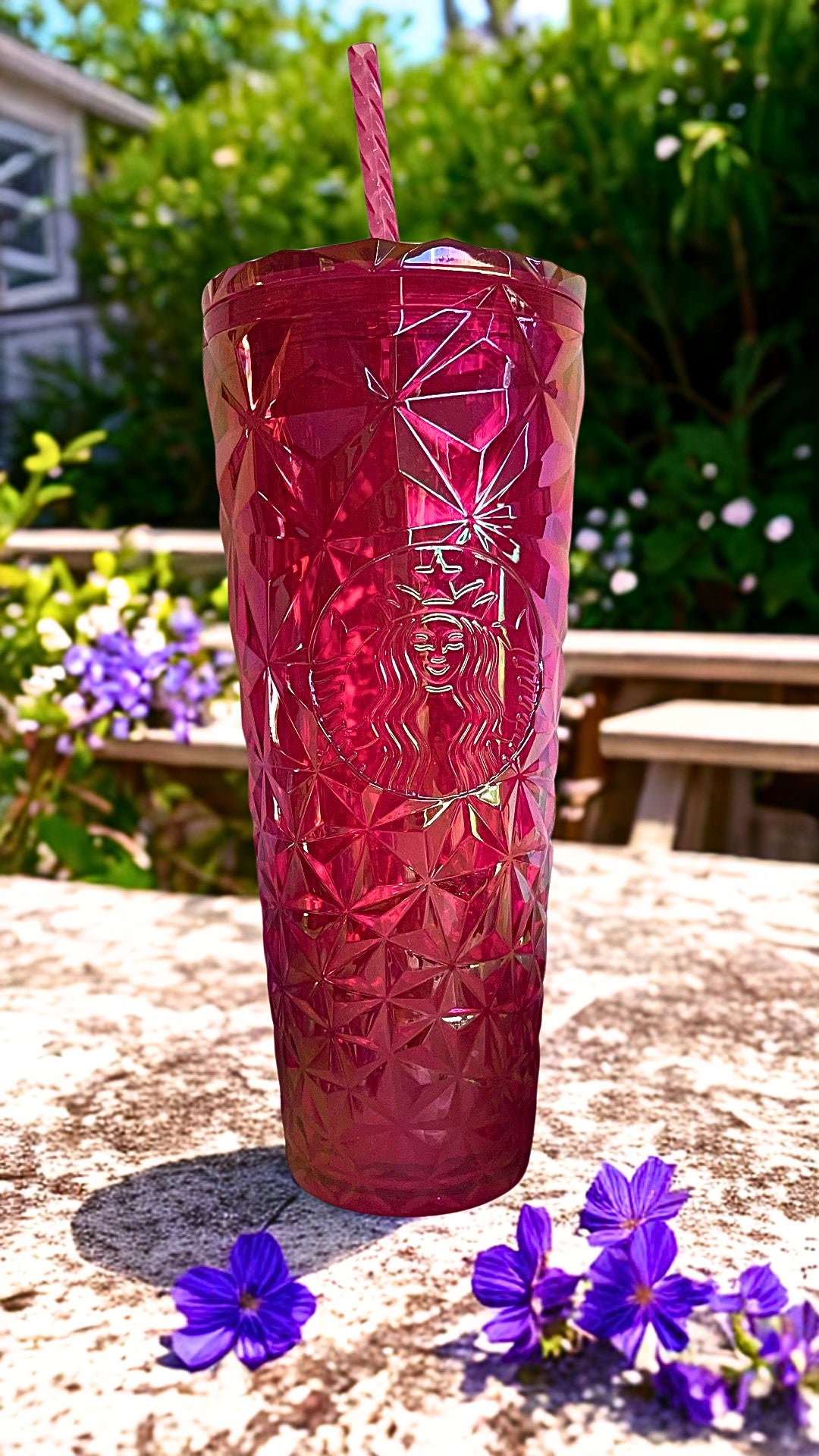 A red Starbucks tumbler with a diamond-cut pattern and a pink striped straw, set outdoors on a stone surface with purple flowers and green foliage in the background.