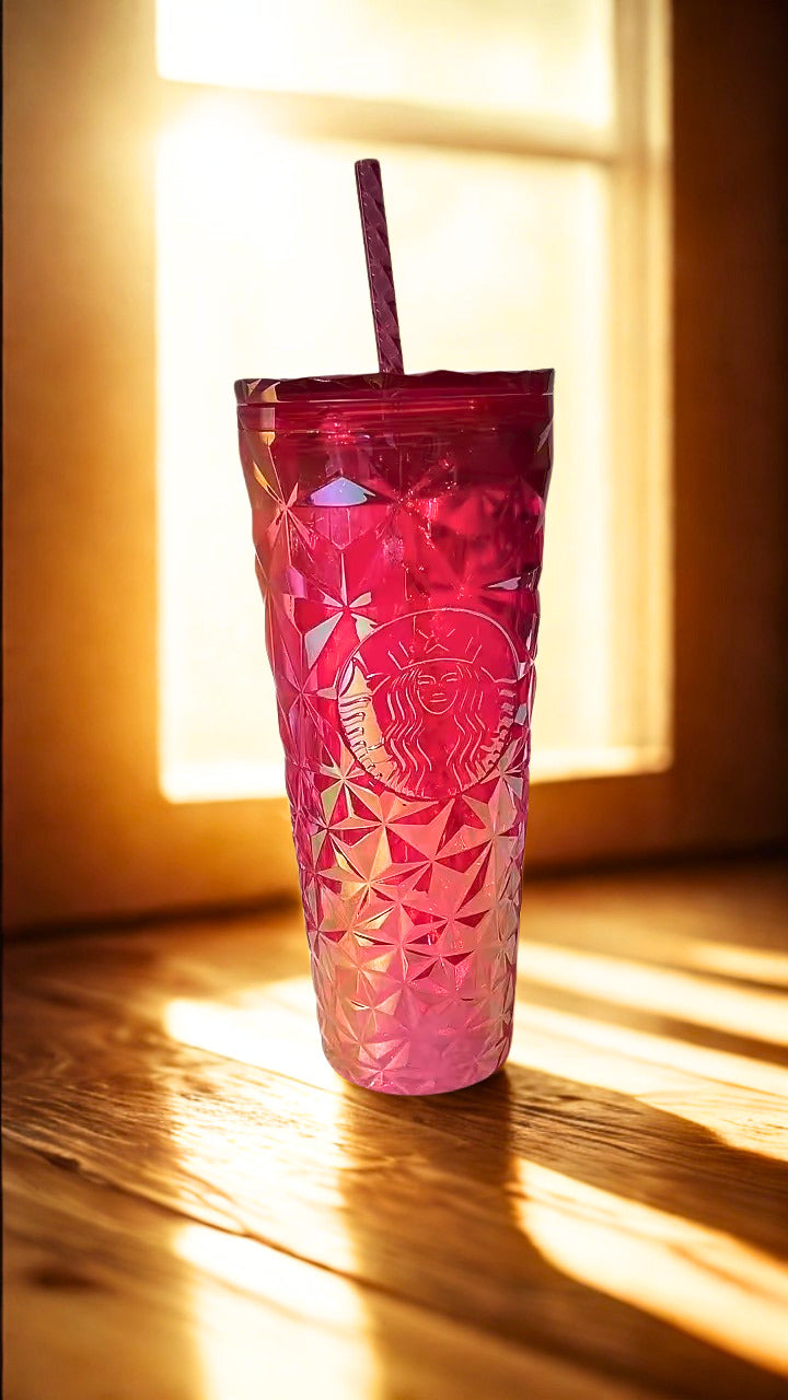 A tall, translucent pink Starbucks  tumbler with a faceted geometric pattern and a dark pink straw, placed on a wooden surface in warm sunlight near a window.