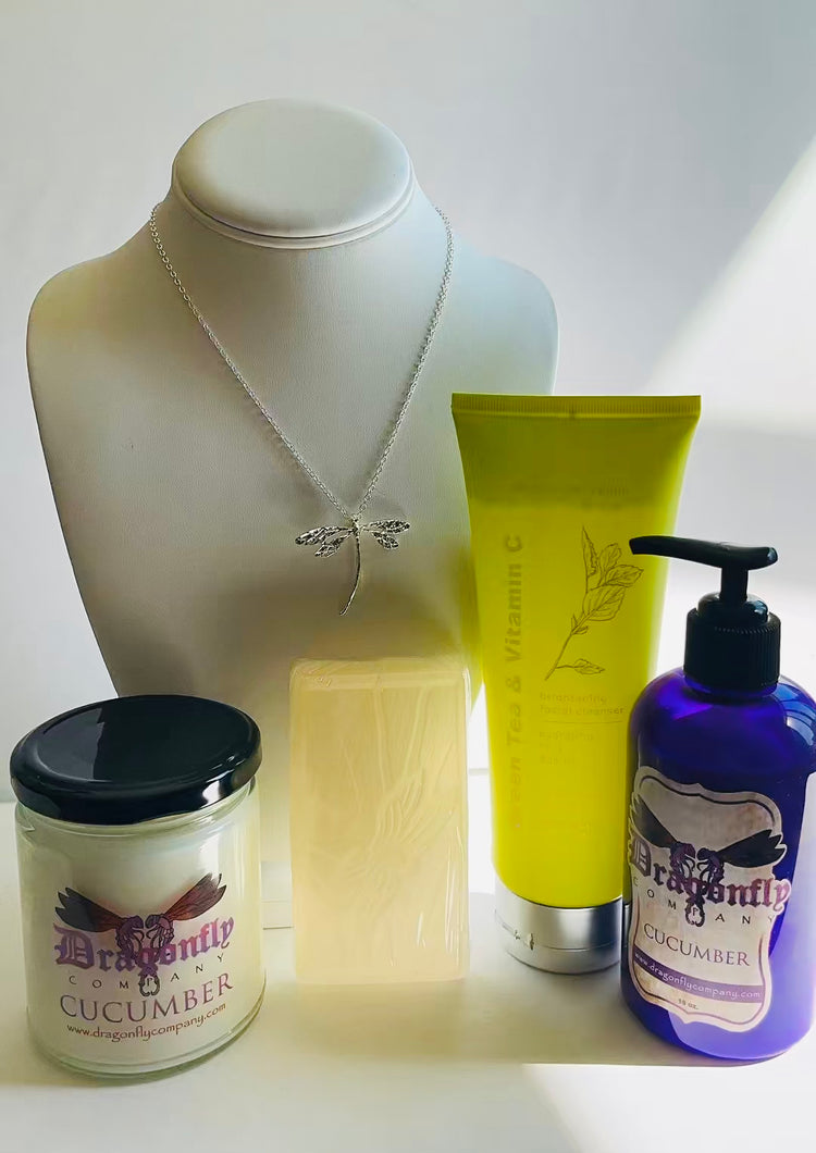 A curated collection of wellness and beauty products including a silver dragonfly necklace ,a Green Tea & Vitamin C face wash, a purple pump bottle of Cucumber lotion, a candle  and a bar of soap, arranged on a white surface.