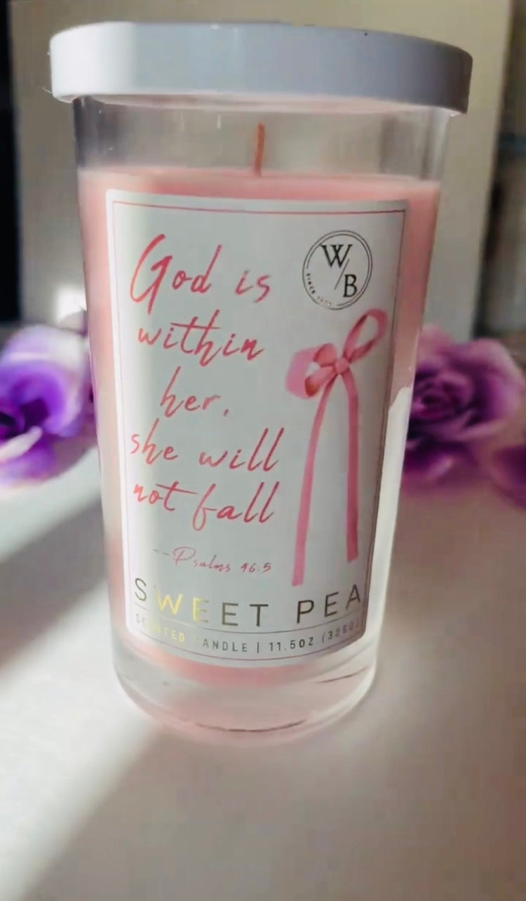 God is within her Candle in a glass jar with text and branding, placed on a surface with flowers in the background.
