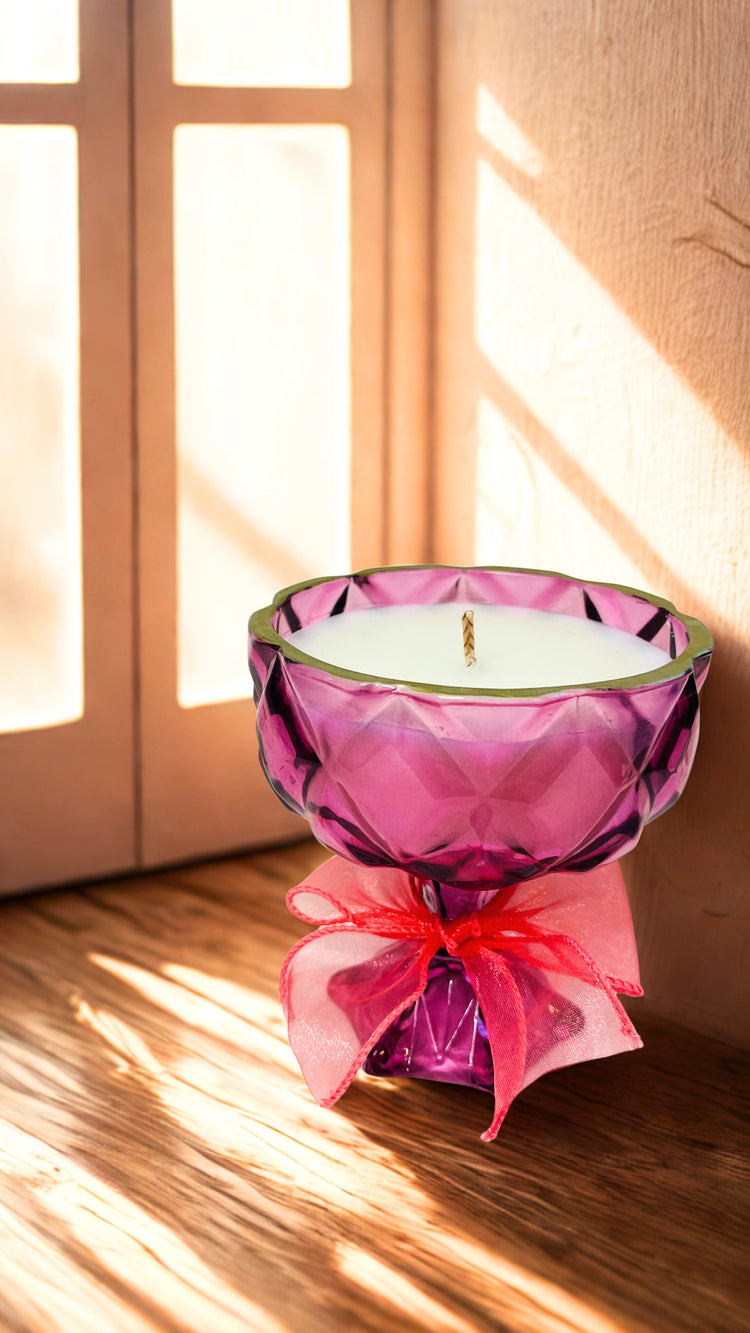 Pink glass candle holder with a pink bow on a wooden surface, sunlight streaming through a window. 