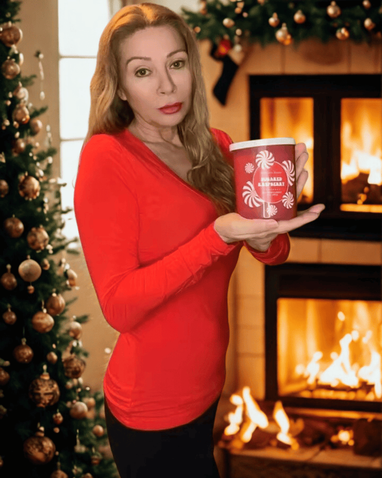 Woman in red shirt holding sugared raspberry two wick candle front of a fireplace and Christmas tree
