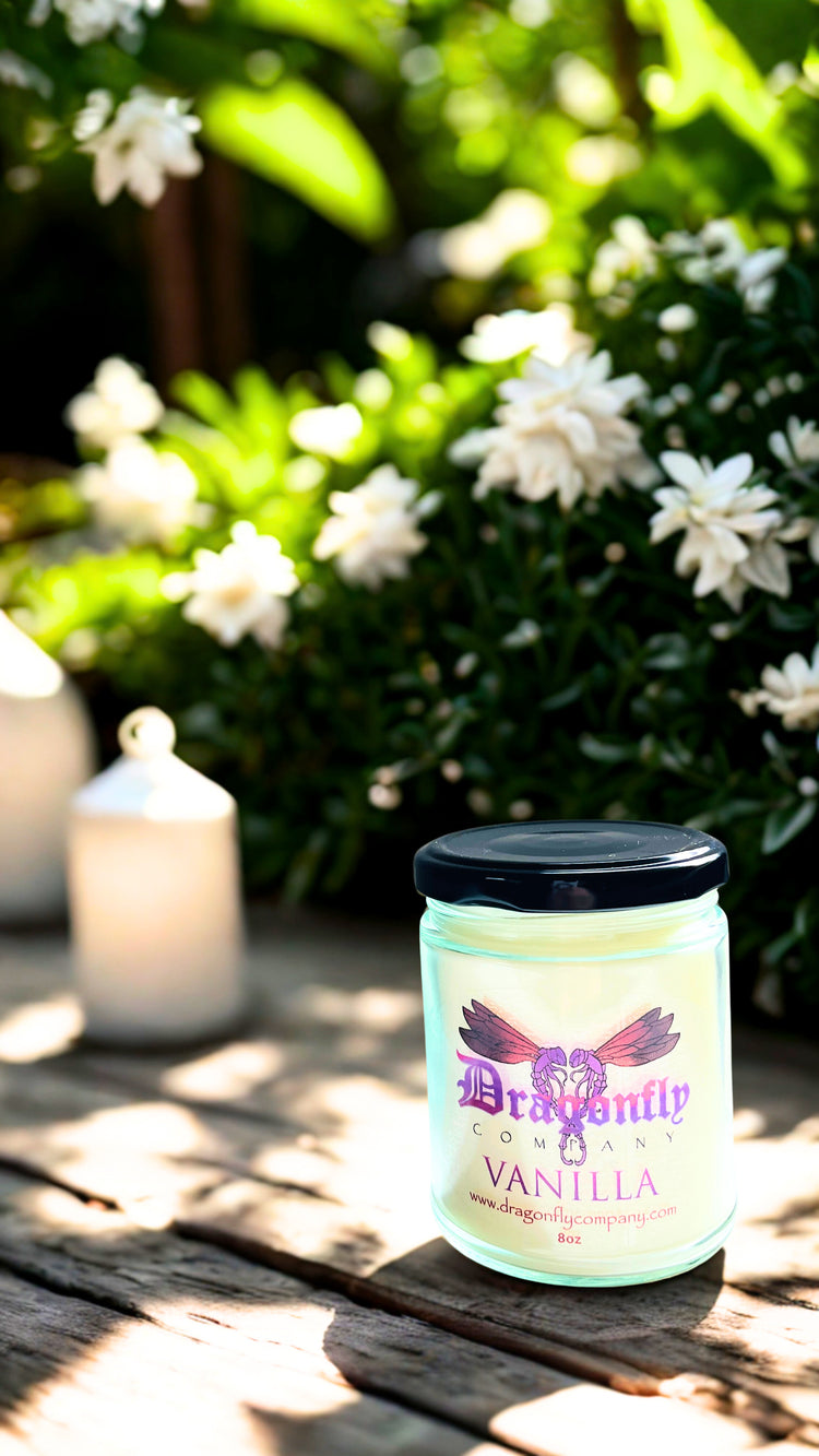 Jar of Vanilla candle with a dragonfly logo on a wooden surface with flowers in the background